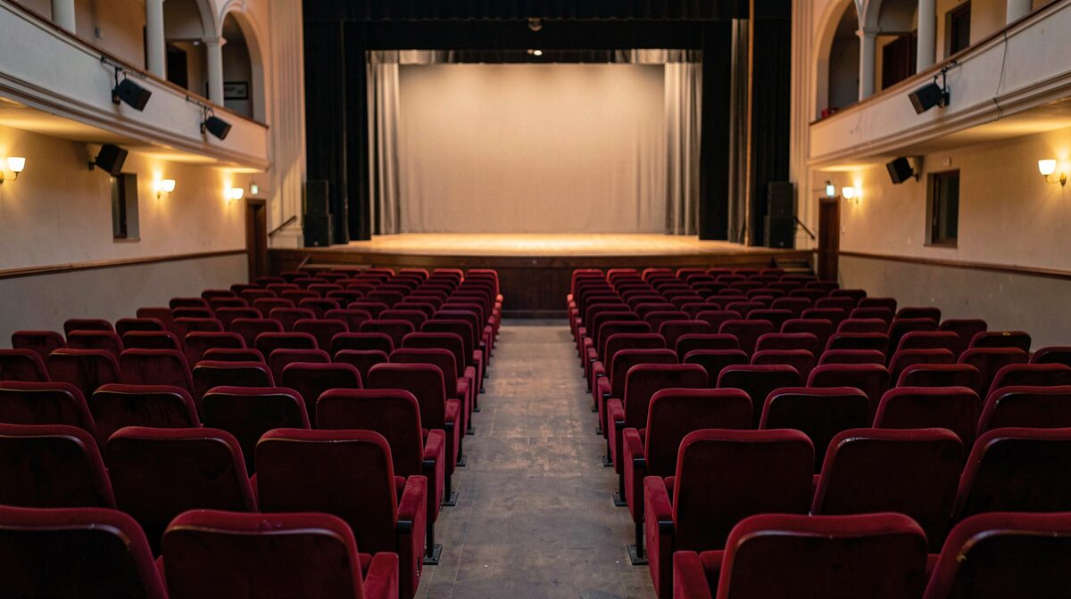An empty small town theater auditorium at dusk, red velvet seats facing a lit stage, framed from the back row with warm house lights visible.