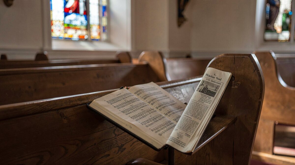 A quiet church pew with an open hymnal and a printed weekly bulletin, lit by soft morning light through a stained glass window, photographed in close framing from the side.