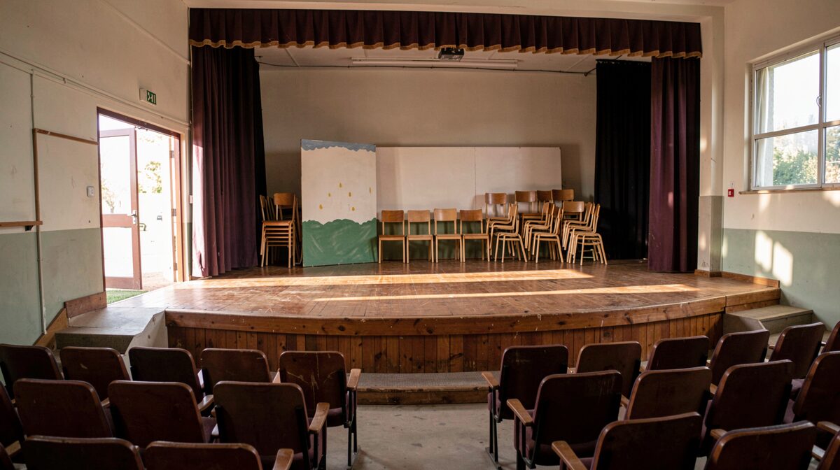 A school auditorium stage with simple painted scenery and stacked chairs, late afternoon light through exit doors, wide shot from the empty seats.